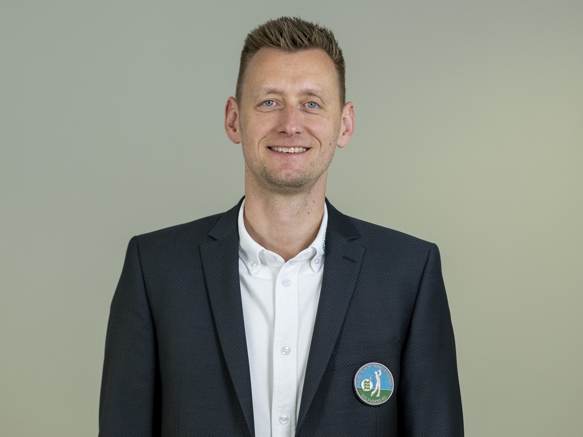 Portrait vor demselben grauen Hintergrund: zweiter Mann im Sakko mit Verbandswappen