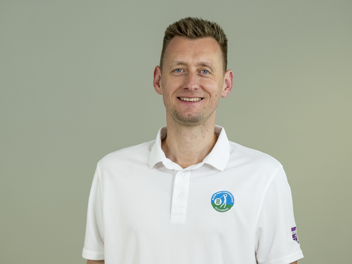Portrait vor demselben grauen Hintergrund: Mann im weißen Polo mit Verbandswappen