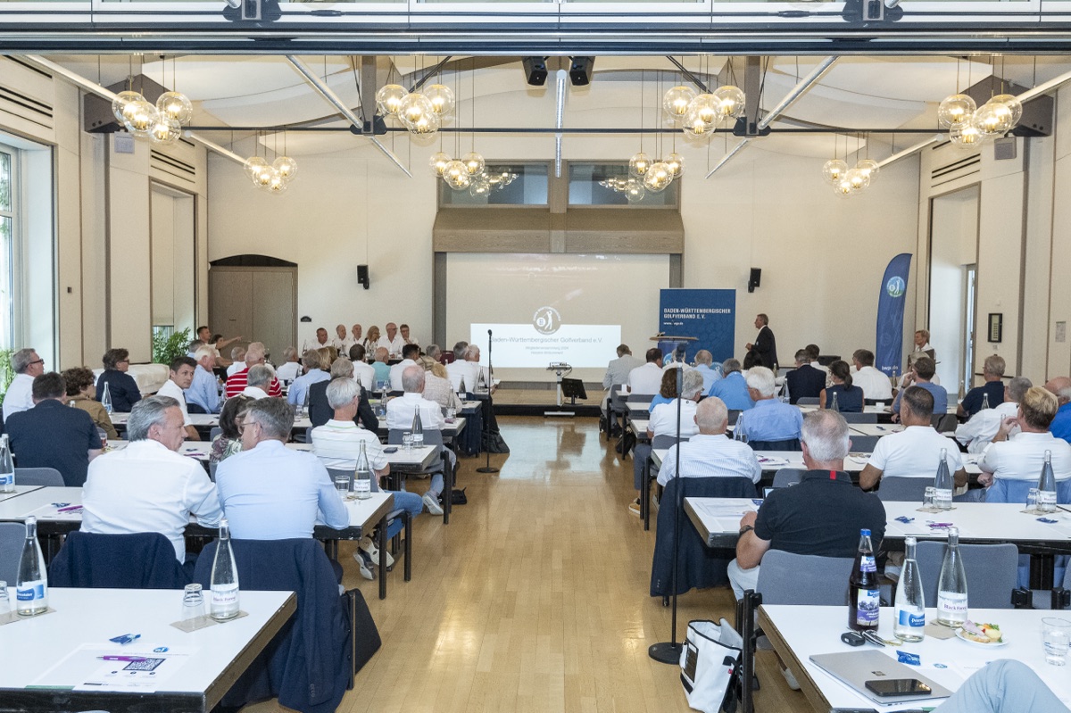 Mitgliederversammlung 2024: Saal mit Tischreihen, Podium und Leinwand