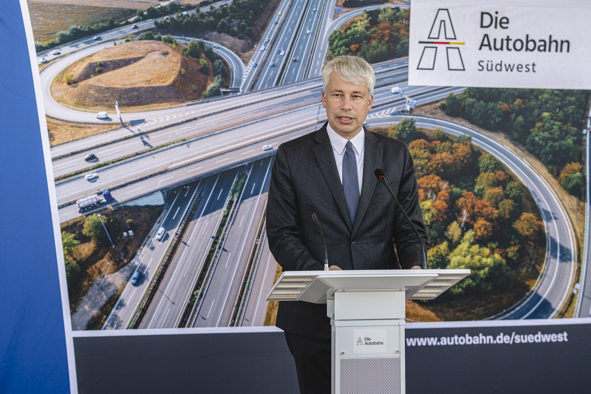 Redner am Pult vor einer Luftaufnahme eines Autobahnkreuzes